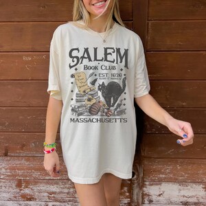 May include: Cream-colored t-shirt with a Salem Book Club graphic. The design includes a black cat, pumpkins, books, and the text "EST 1626" and "Massachusetts". The shirt has short sleeves and a relaxed fit.