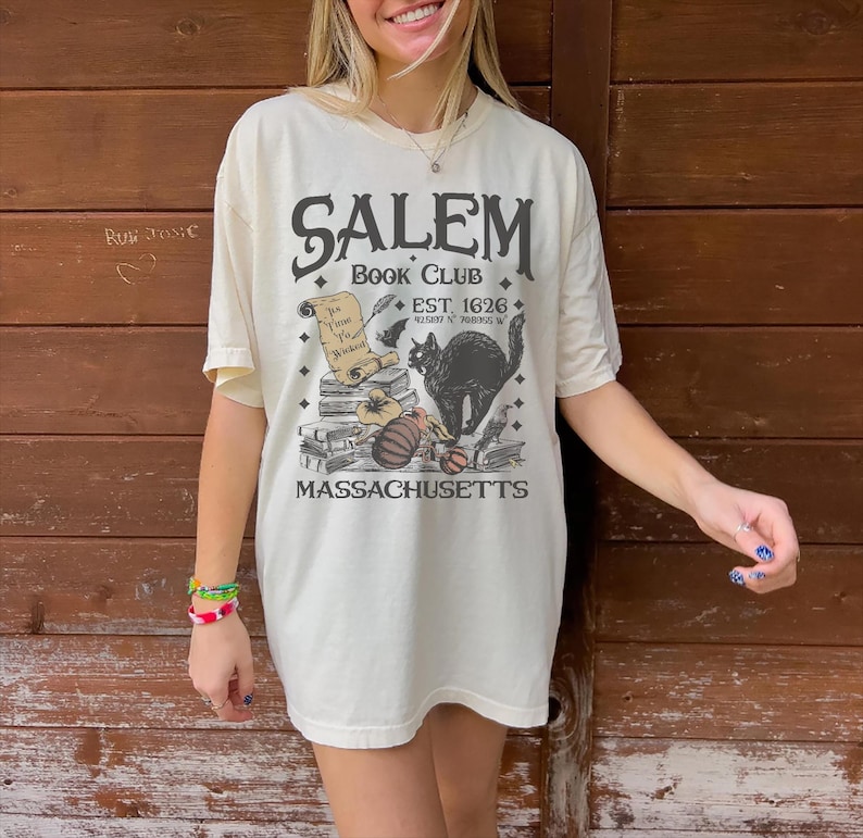 May include: Cream-colored t-shirt with a Salem Book Club graphic. The design includes a black cat, pumpkins, books, and the text "EST 1626" and "Massachusetts". The shirt has short sleeves and a relaxed fit.