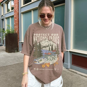 May include: A brown t-shirt with a graphic print of a mountain scene and a fish. The text on the shirt reads "Protect Our National Parks" and "Explore Responsibly".