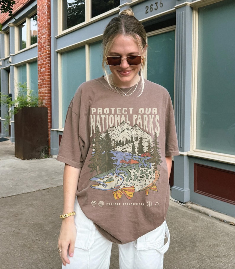 May include: A brown t-shirt with a graphic print of a mountain scene and a fish. The text on the shirt reads "Protect Our National Parks" and "Explore Responsibly".