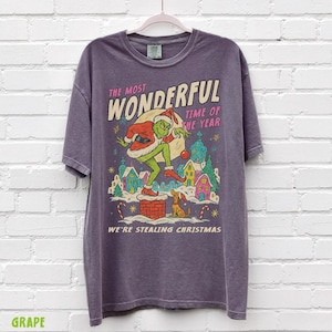 Puede incluir: Camiseta morada con un dise&ntilde;o tem&aacute;tico de Grinch. El dise&ntilde;o incluye el texto "The Most Wonderful Time of the Year" y "We're Stealing Christmas". El Grinch est&aacute; representado con un traje de Pap&aacute; Noel, con un perro y casas coloridas.