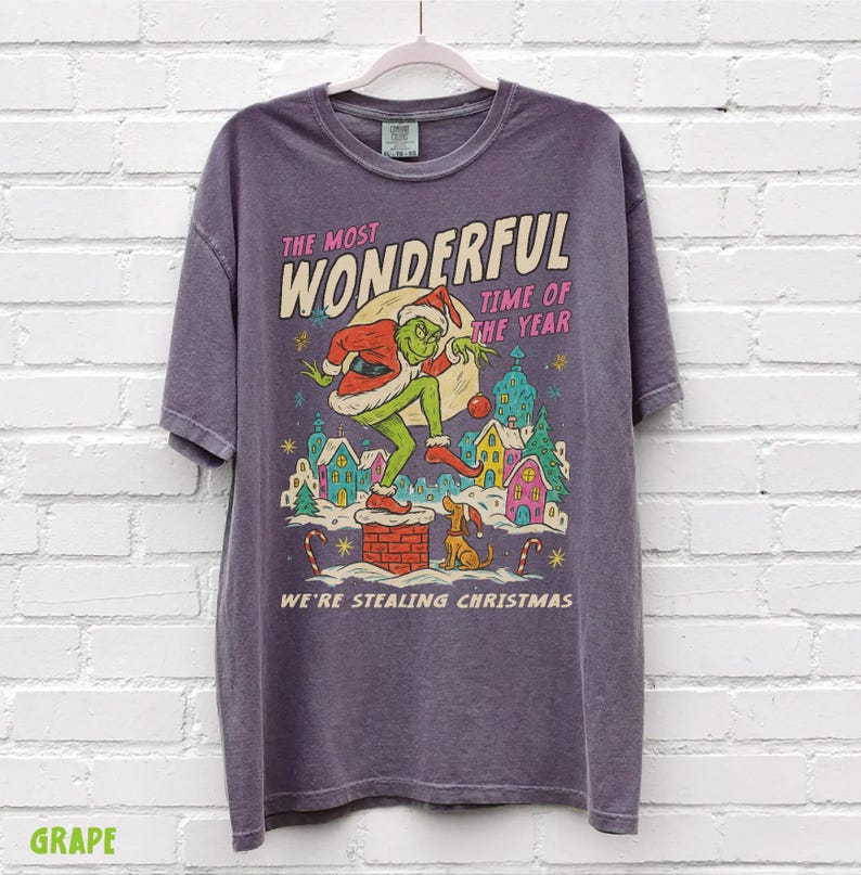 Puede incluir: Camiseta morada con un dise&ntilde;o tem&aacute;tico de Grinch. El dise&ntilde;o incluye el texto "The Most Wonderful Time of the Year" y "We're Stealing Christmas". El Grinch est&aacute; representado con un traje de Pap&aacute; Noel, con un perro y casas coloridas.