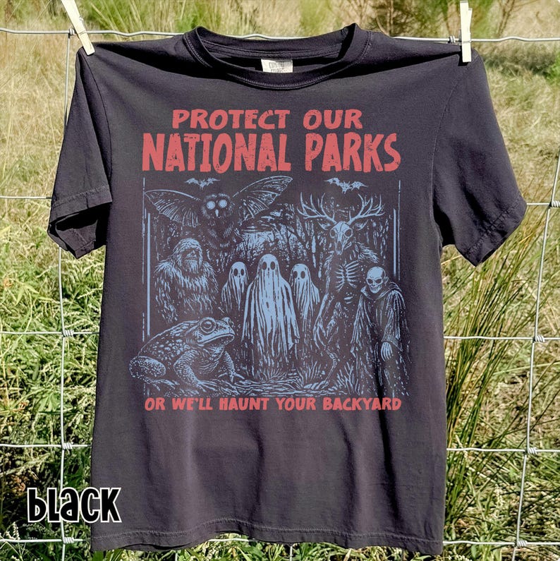 May include: A black short-sleeve t-shirt featuring a graphic print with the red text "PROTECT OUR NATIONAL PARKS" and "OR WE'LL HAUNT YOUR BACKYARD". The illustration depicts a toad, a yeti, an owl, three ghosts, and a deer skull creature.