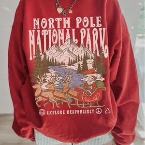 May include: Red sweatshirt with a graphic of Santa Claus in a sleigh pulled by reindeer, with the text "NORTH POLE NATIONAL PARK" and "EXPLORE RESPONSIBLY". The design includes mountains, trees, and a lake.
