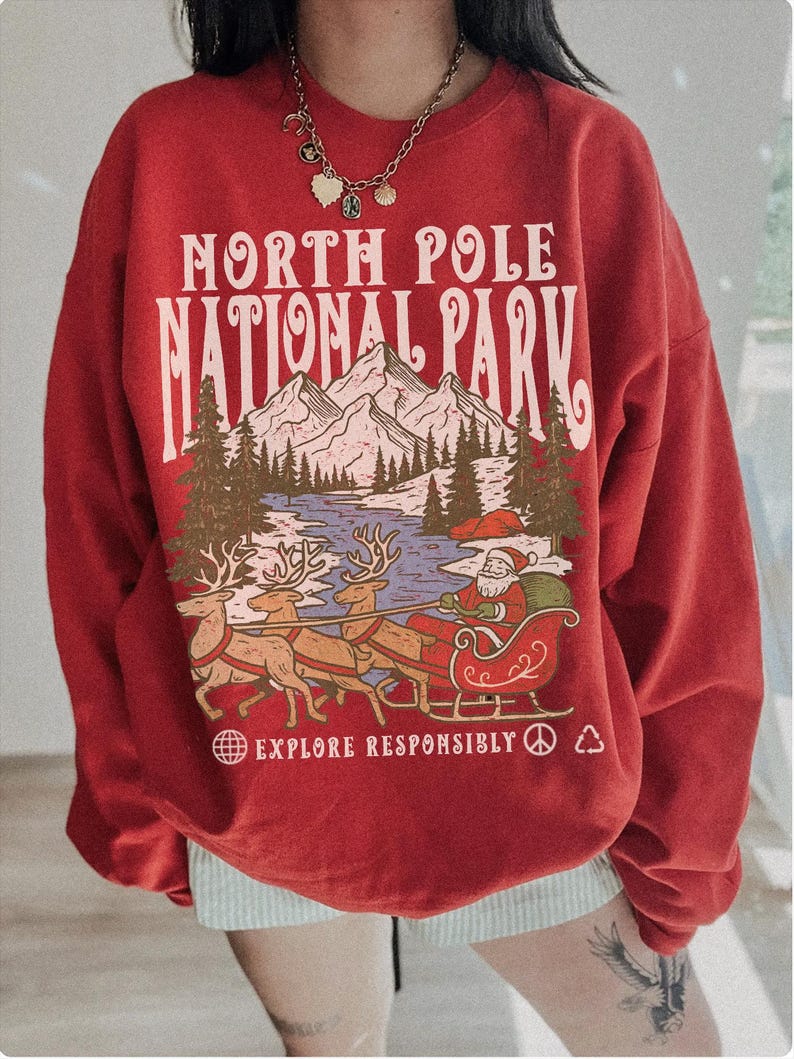 May include: Red sweatshirt with a graphic of Santa Claus in a sleigh pulled by reindeer, with the text "NORTH POLE NATIONAL PARK" and "EXPLORE RESPONSIBLY". The design includes mountains, trees, and a lake.