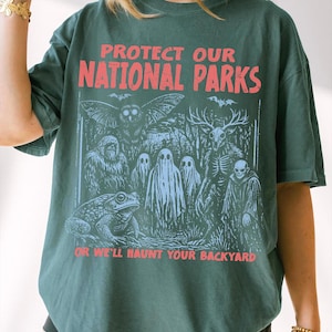 May include: A dark green t-shirt featuring the red text PROTECT OUR NATIONAL PARKS at the top and OR WE'LL HAUNT YOUR BACKYARD at the bottom. The graphic depicts a forest scene with an owl, a sasquatch, ghosts, a deer skull creature, and a toad.