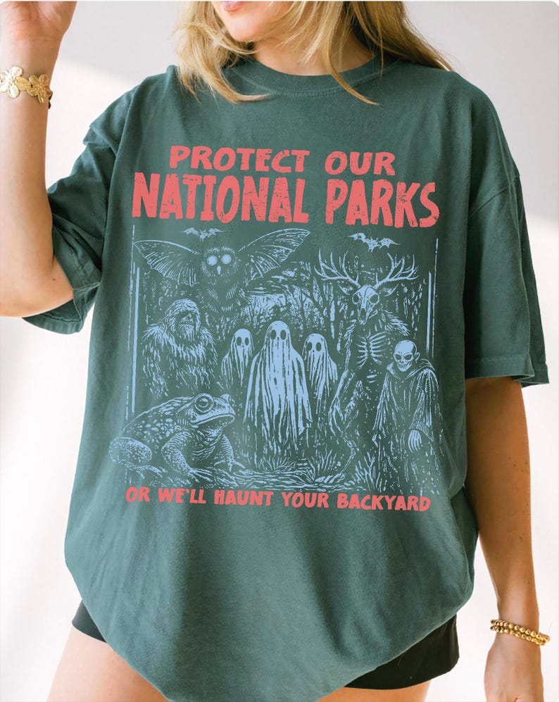 May include: A dark green t-shirt featuring the red text PROTECT OUR NATIONAL PARKS at the top and OR WE'LL HAUNT YOUR BACKYARD at the bottom. The graphic depicts a forest scene with an owl, a sasquatch, ghosts, a deer skull creature, and a toad.