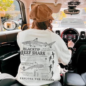 May include: Cream-coloured t-shirt with a black and white graphic of a blacktip reef shark and scientific details. The text reads "BLACKTIP REEF SHARK" and "EXPLORE THE OCEANS". The design includes shark illustrations and facts about the species.