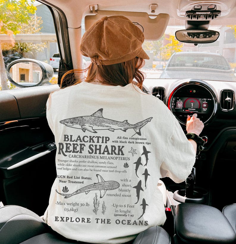 May include: Cream-coloured t-shirt with a black and white graphic of a blacktip reef shark and scientific details. The text reads "BLACKTIP REEF SHARK" and "EXPLORE THE OCEANS". The design includes shark illustrations and facts about the species.