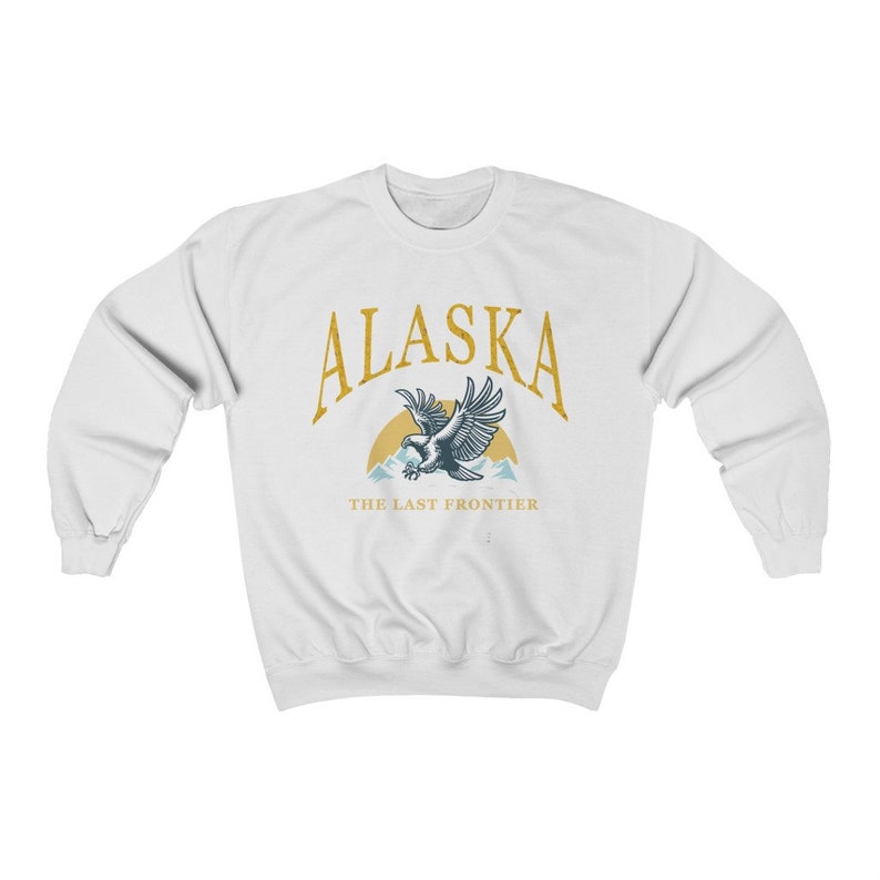 Peut inclure: Sweat-shirt blanc avec un graphique dor&eacute; d'un aigle volant au-dessus des montagnes avec le texte "Alaska The Last Frontier".