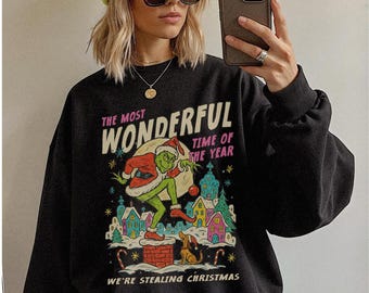 Può includere: Felpa nera con grafica del Grinch vestito da Babbo Natale, con la scritta "THE MOST WONDERFUL TIME OF THE YEAR" e "WE'RE STEALING CHRISTMAS". Il design include un villaggio festivo.