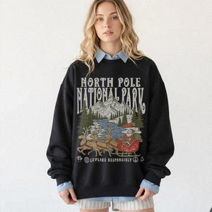 May include: Black sweatshirt with a graphic featuring Santa Claus in a sleigh pulled by reindeer, with the text "NORTH POLE NATIONAL PARK" and "EXPLORE RESPONSIBLY". The design includes mountains, trees, and a river.