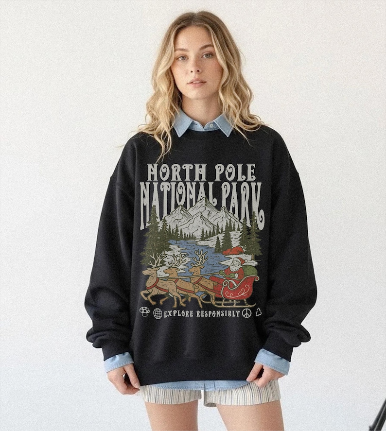 May include: Black sweatshirt with a graphic featuring Santa Claus in a sleigh pulled by reindeer, with the text "NORTH POLE NATIONAL PARK" and "EXPLORE RESPONSIBLY". The design includes mountains, trees, and a river.