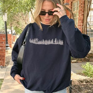 Op de afbeelding: Een marineblauw sweatshirt met lange mouwen en een ronde hals, voorzien van een lichtgrijze grafische print van een boslandschap met diverse dennenbomen op de borst. Het sweatshirt heeft een relaxte pasvorm met geribbelde boorden en zoom.