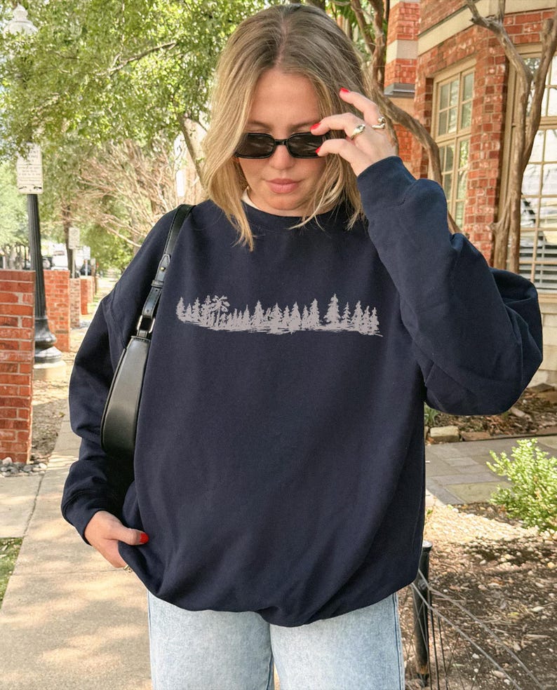 Op de afbeelding: Een marineblauw sweatshirt met lange mouwen en een ronde hals, voorzien van een lichtgrijze grafische print van een boslandschap met diverse dennenbomen op de borst. Het sweatshirt heeft een relaxte pasvorm met geribbelde boorden en zoom.