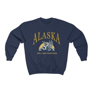 Peut inclure: Sweat-shirt bleu marine avec un graphique d'un aigle chauve volant au-dessus des montagnes avec le texte "Alaska The Last Frontier" en jaune.