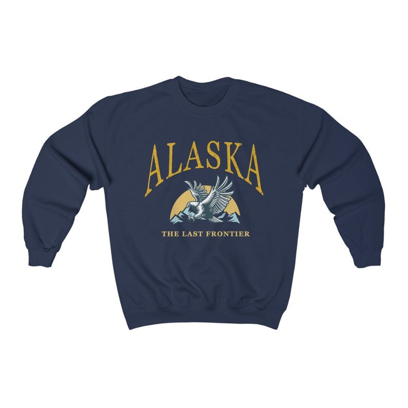 Peut inclure: Sweat-shirt bleu marine avec un graphique d'un aigle chauve volant au-dessus des montagnes avec le texte "Alaska The Last Frontier" en jaune.