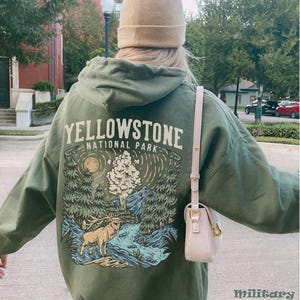 May include: A green hooded sweatshirt with a graphic print of a forest scene with a deer, a river, and a geyser. The text "Yellowstone National Park" is printed on the front of the sweatshirt.