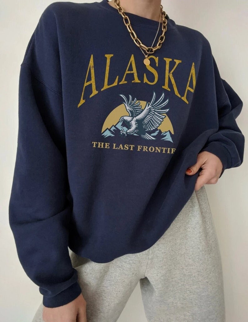 Peut inclure: Un sweat-shirt bleu marine avec un imprim&eacute; graphique d'un aigle volant au-dessus des montagnes. Le texte "ALASKA THE LAST FRONTIF" est imprim&eacute; sur le sweat-shirt.
