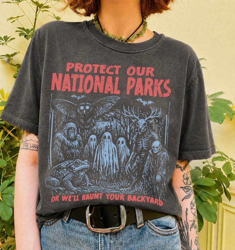 May include: A charcoal gray short-sleeve t-shirt featuring a graphic print in red and blue. The text reads PROTECT OUR NATIONAL PARKS OR WE'LL HAUNT YOUR BACKYARD. The illustration depicts an owl, a sasquatch, ghosts, a deer skull, and a toad.