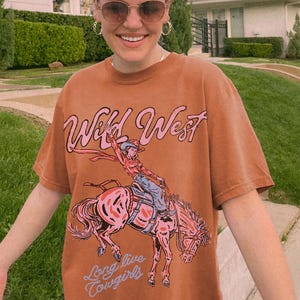 Puede incluir: Una camiseta de manga corta color &oacute;xido con un estampado gr&aacute;fico de una persona montando a caballo. El texto "Wild West" aparece en rosa sobre el gr&aacute;fico, y el texto "Long live cowgirls" aparece en azul claro debajo de la ilustraci&oacute;n.