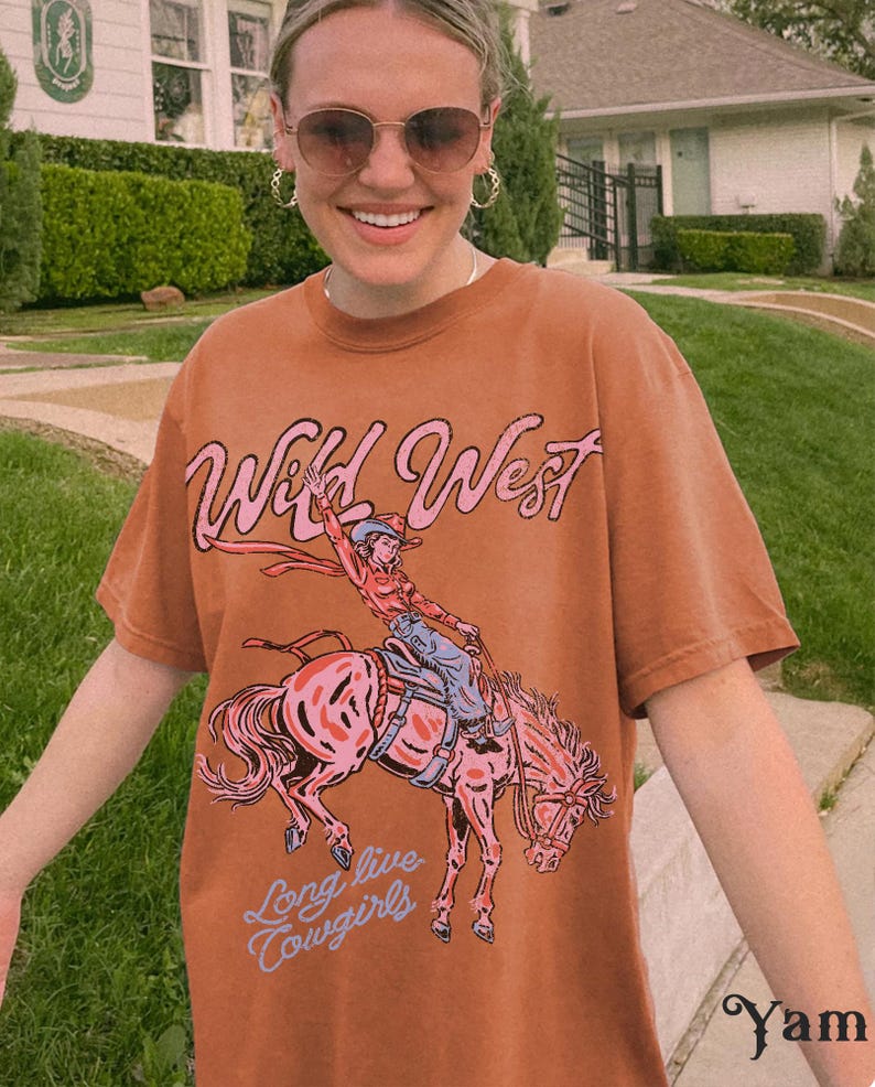Puede incluir: Una camiseta de manga corta color &oacute;xido con un estampado gr&aacute;fico de una persona montando a caballo. El texto "Wild West" aparece en rosa sobre el gr&aacute;fico, y el texto "Long live cowgirls" aparece en azul claro debajo de la ilustraci&oacute;n.