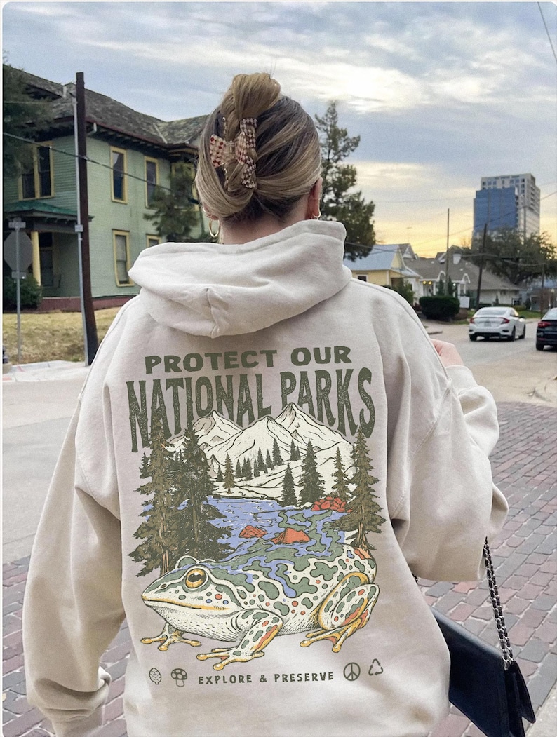 May include: A beige hooded sweatshirt featuring a graphic on the back with the text PROTECT OUR NATIONAL PARKS and EXPLORE & PRESERVE. The illustration depicts a large frog in the foreground with a mountain, forest, and lake scene behind it.