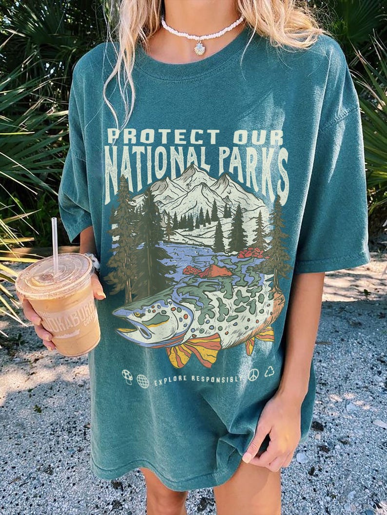 May include: A teal green t-shirt with a graphic print of a mountain scene and a fish. The text on the shirt reads "Protect Our National Parks" and "Explore Responsibly".