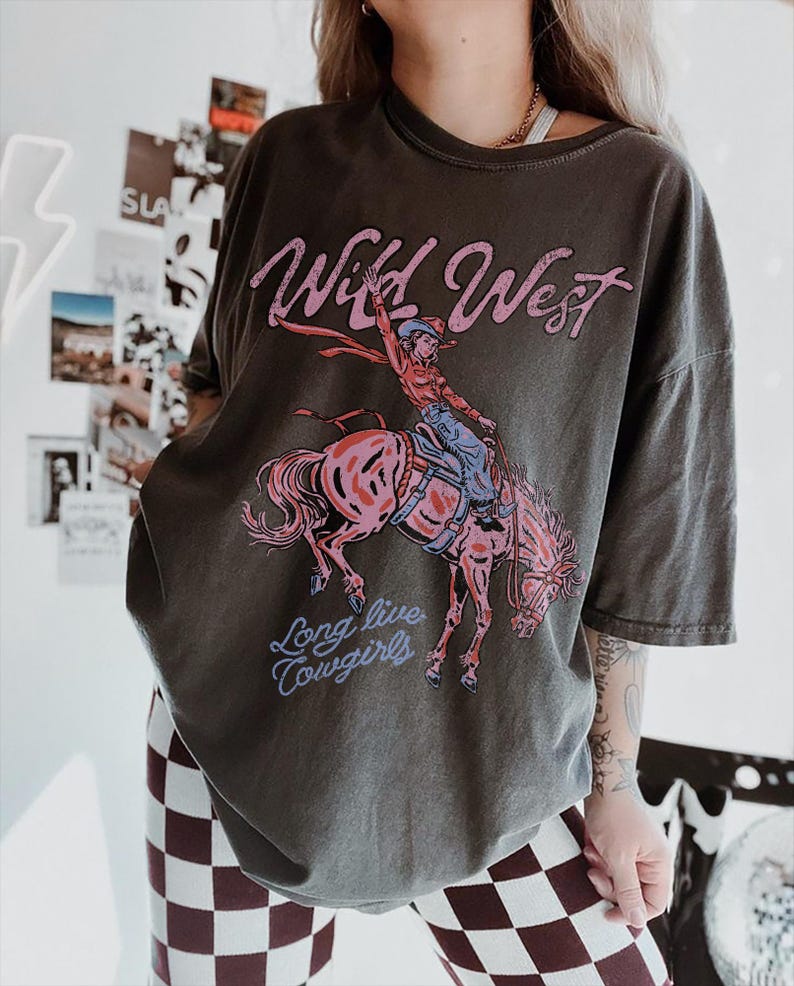 Puede incluir: Una camiseta gris carb&oacute;n de corte oversize con un estampado gr&aacute;fico de una vaquera montando un caballo encabritado. El texto "Wild West" aparece en rosa sobre la ilustraci&oacute;n, y "Long live cowgirls" est&aacute; impreso en azul bajo el caballo.