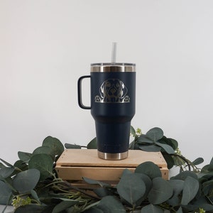 May include: A navy blue stainless steel travel mug with a black dog silhouette engraved on the side. The mug has a lid and a handle. It is sitting on a wooden crate with green leaves around it.