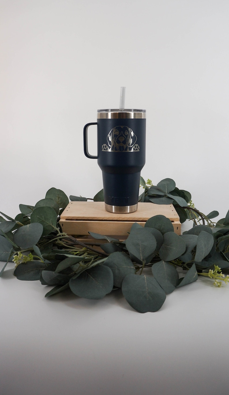 May include: A navy blue stainless steel travel mug with a black dog silhouette engraved on the side. The mug has a lid and a handle. It is sitting on a wooden crate with green leaves around it.