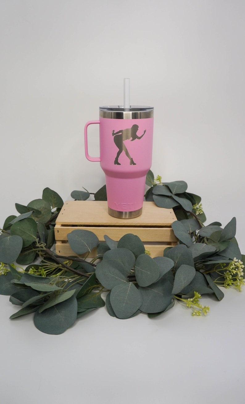 May include: Pink stainless steel travel mug with a black silhouette of a person on it. The mug has a handle and a straw. It is sitting on a wooden crate.