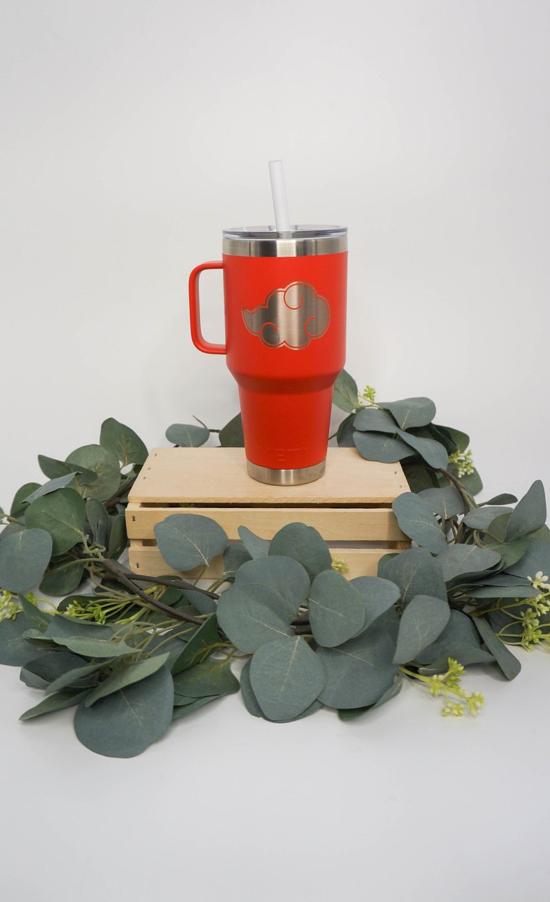 May include: Red stainless steel travel mug with a handle and a straw. The mug has a silver cloud design on the front. The mug is sitting on a wooden crate surrounded by green leaves.