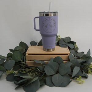 May include: A purple Yeti tumbler with a handle and a straw. The tumbler has a logo that says "SoCal Equine" with a horse silhouette and a heartbeat graphic. The tumbler is sitting on a wooden crate with green leaves around it.