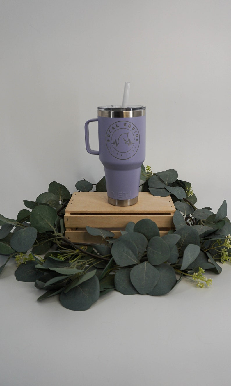 May include: A purple Yeti tumbler with a handle and a straw. The tumbler has a logo that says "SoCal Equine" with a horse silhouette and a heartbeat graphic. The tumbler is sitting on a wooden crate with green leaves around it.