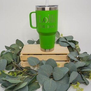 May include: A green travel mug with a handle and a straw. The mug has the name "Lolli" and the text "Est. 2019" engraved on it. The mug is sitting on a wooden crate surrounded by green eucalyptus leaves.