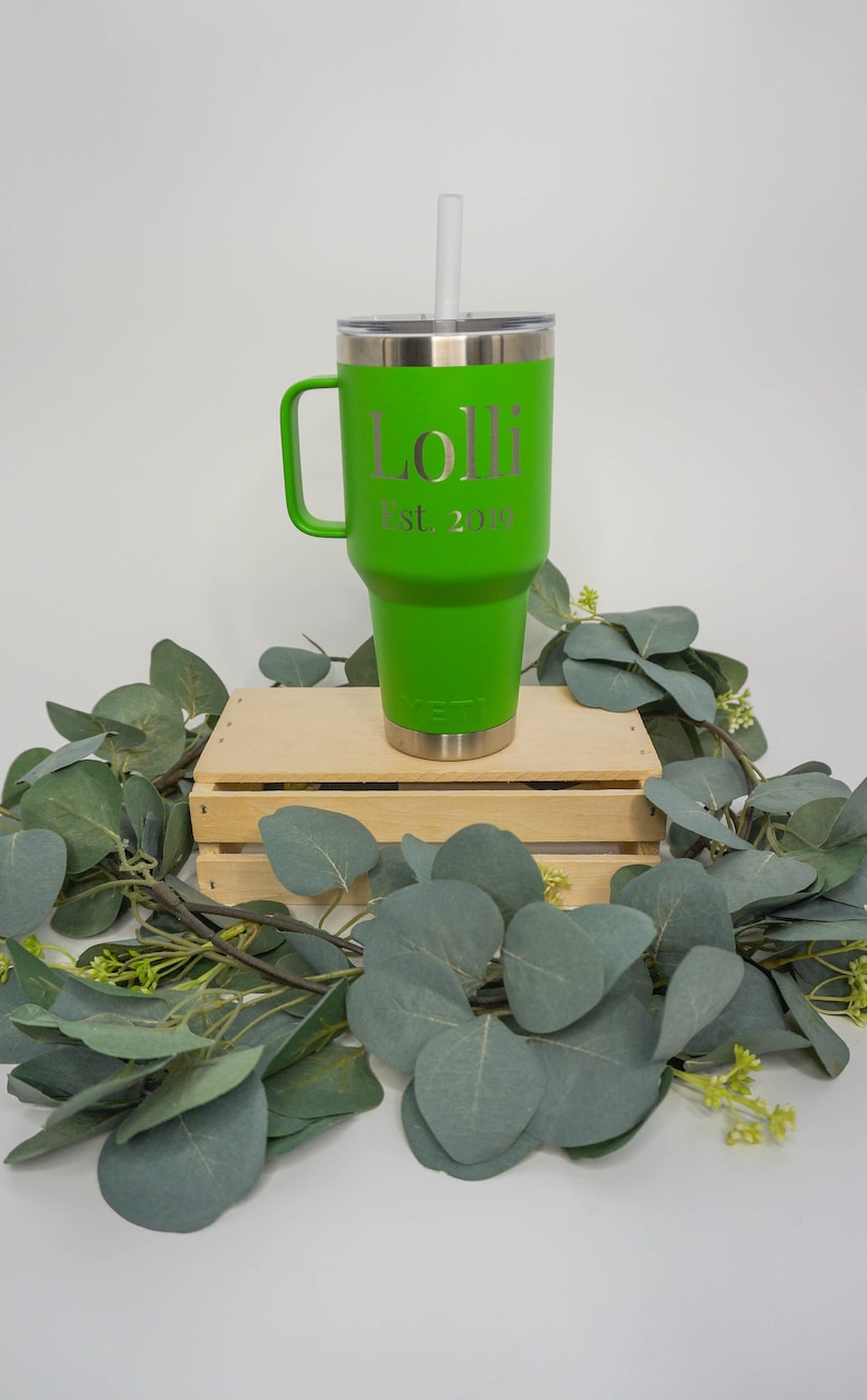 May include: A green travel mug with a handle and a straw. The mug has the name "Lolli" and the text "Est. 2019" engraved on it. The mug is sitting on a wooden crate surrounded by green eucalyptus leaves.