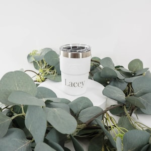 May include: A white insulated tumbler with a silver rim and a clear lid. The name "Lacey" is printed in a gray script on the front of the tumbler. The tumbler is surrounded by green eucalyptus leaves and sits on a white platform.