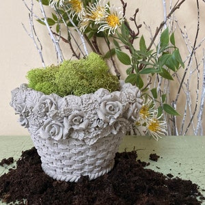 May include: A gray concrete planter shaped like a basket with a floral design around the top. The planter is filled with green moss and is sitting on a bed of dark brown soil.