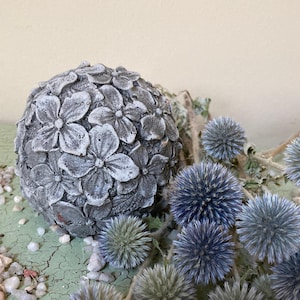 May include: A gray concrete sphere with a floral design, surrounded by blue thistle flowers on a green surface.