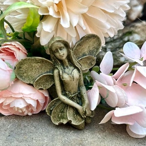 May include: A small, detailed figurine of a fairy with wings, wearing a dress and holding her hands together. The fairy is surrounded by pink and white flowers.