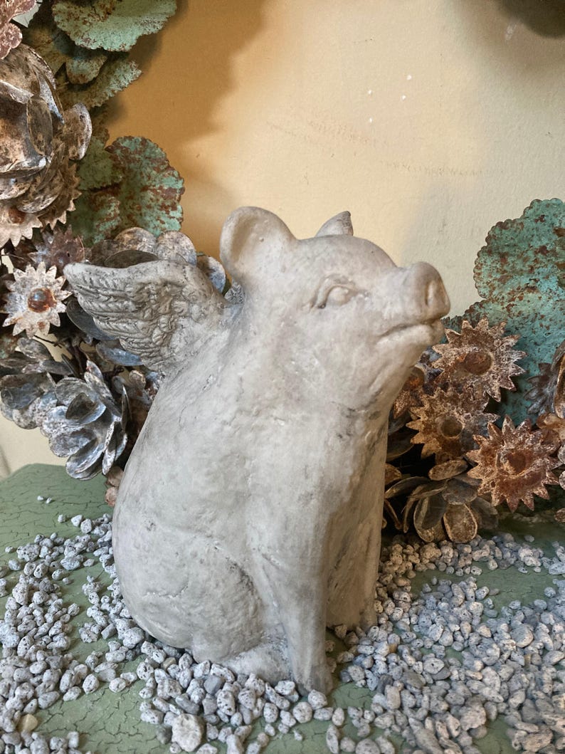 side profile of flying pig garden statue sitting on a green table displayed with various grey stones and a metal floral farmhouse wreath