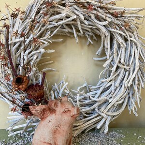 side profile of flying pig garden statue in Terra cotta coloring sitting on a green table displayed with various grey stones and a white branch wreath