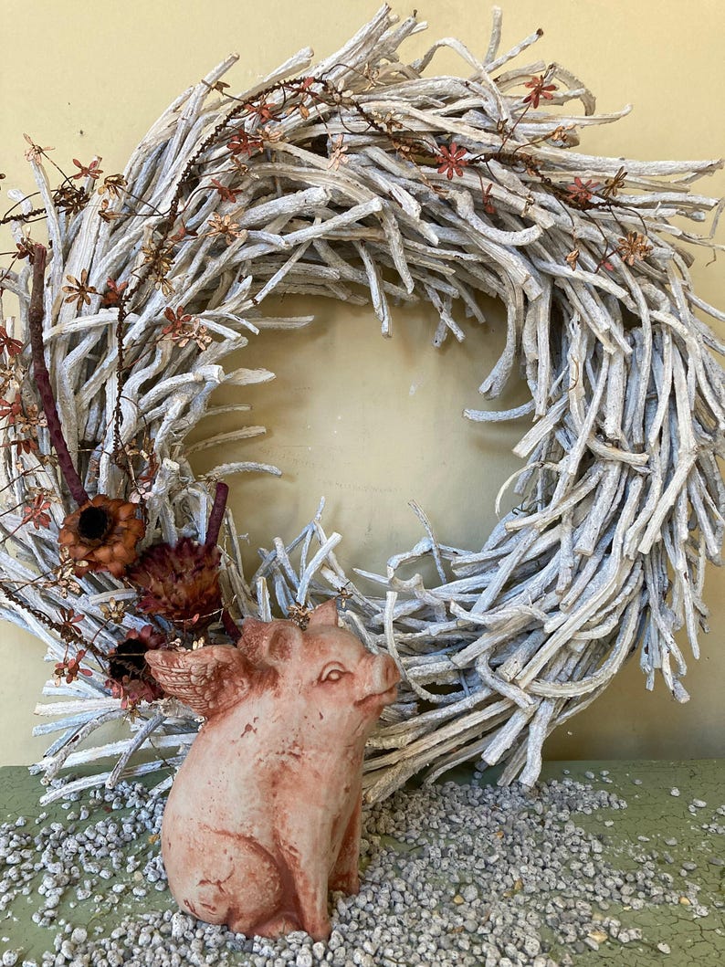 side profile of flying pig garden statue in Terra cotta coloring sitting on a green table displayed with various grey stones and a white branch wreath