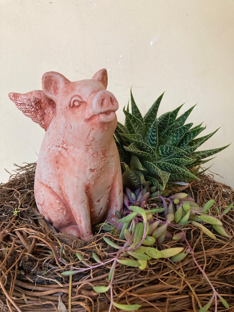 flying pig garden statue in Terra cotta color nestled in a garden sitting atop a nest of small branches surround by green succulents and a large agave plant