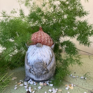 May include: A small gray gnome figurine with a red and brown acorn hat. The gnome is holding a small brown acorn in its hands. The gnome is sitting on a green surface with white pebbles scattered around it.