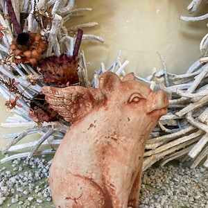 side profile of flying pig garden statue in Terra cotta color sitting on a green table displayed with various grey stones and a metal floral farmhouse wreath