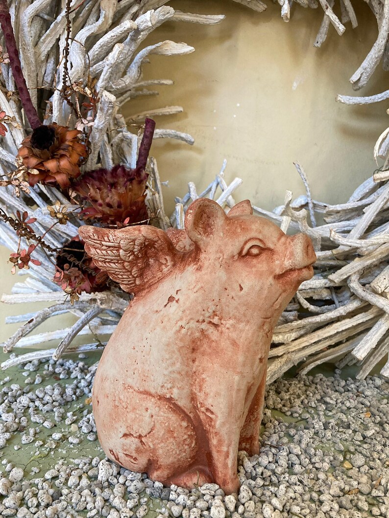 side profile of flying pig garden statue in Terra cotta color sitting on a green table displayed with various grey stones and a metal floral farmhouse wreath