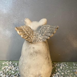 backside wings of flying pig garden statue in stone grey color sitting on a green table displayed with various grey pebbles