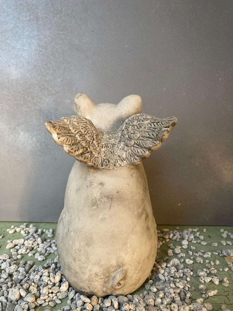 backside wings of flying pig garden statue in stone grey color sitting on a green table displayed with various grey pebbles